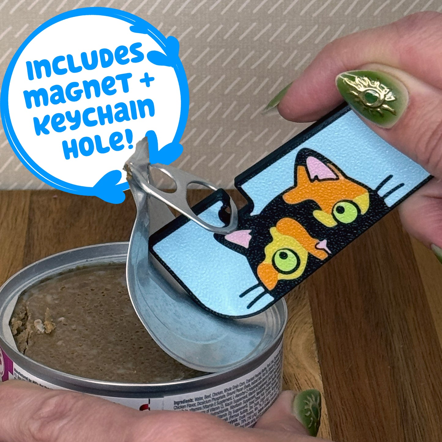 A Tab Buddy XL with a black, orange and yellow tortoiseshell cat design is used to peel back the tab lid on a can of cat food. Text reads "Includes magnet + keychain hole!"