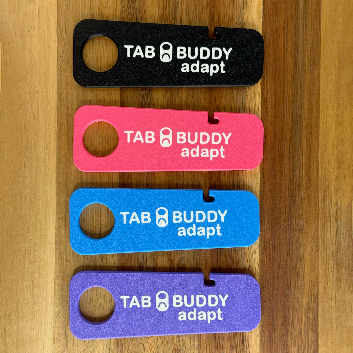Tab Buddy Adapts are shown resting on a wooden surface in four color options: black, bright pink, sky blue and purple.