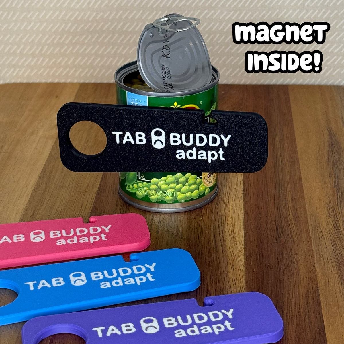 A black Tab Buddy Adapt is magnetized to an open can of peas. Tab Buddy Adapts in pink, blue and purple rest on a wooden surface in the foreground. Text reads "Magnet inside!"