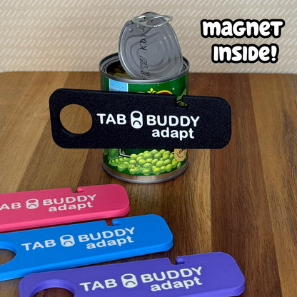 A black Tab Buddy Adapt is magnetized to an open can of peas. Tab Buddy Adapts in pink, blue and purple rest on a wooden surface in the foreground. Text reads "Magnet inside!"