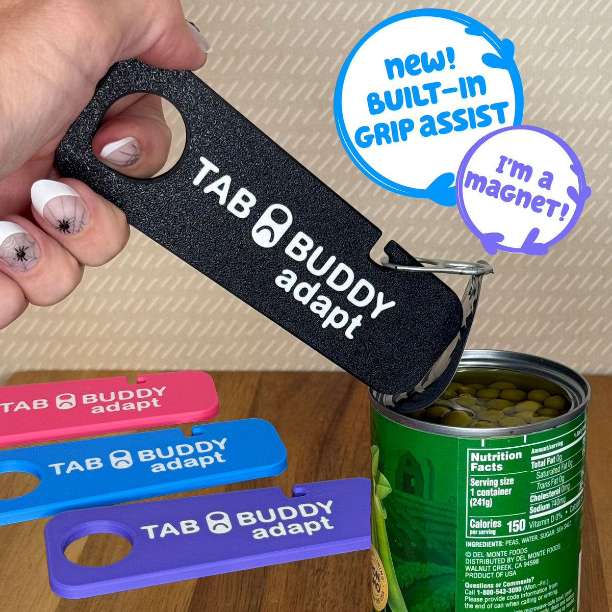 A black Tab Buddy Adapt with a grip assist hole is used to peel back the lid on a can of peas. Pink, blue and purple Tab Buddy adapts are shown resting on the wooden surface in backgroud. Text bubbles read: "New! Built-in grip assist." and "I'm a magnet!"