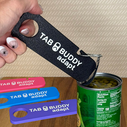 A black Tab Buddy Adapt with a grip assist hole is used to peel back the lid on a can of peas. Pink, blue and purple Tab Buddy adapts are shown resting on the wooden surface in backgroud.