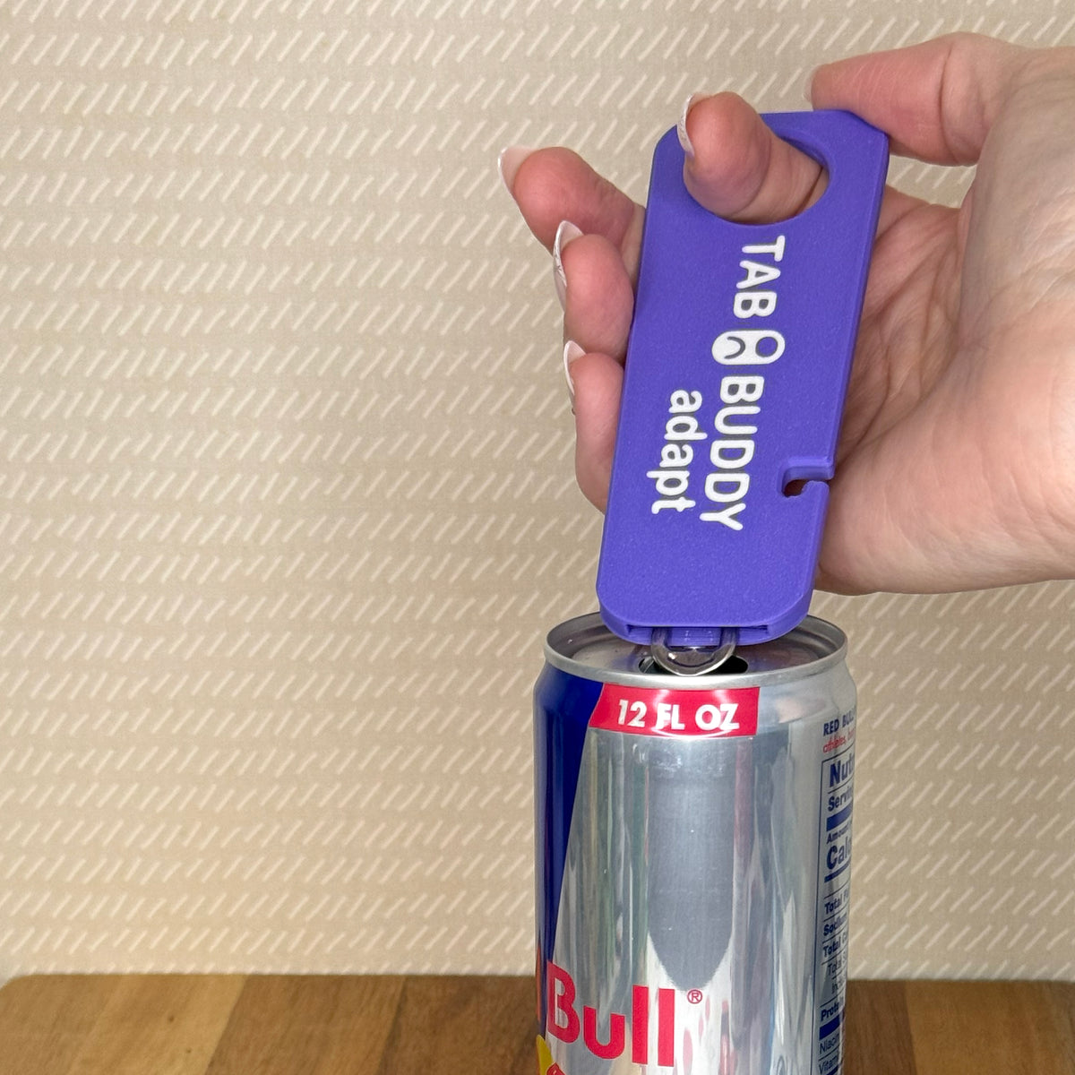 A purple Tab Buddy Adapt is used to open a can of Red Bull. The individual opening the can has placed their pointer finger through the grip assist hole and is lightly palming the Tab Buddy upward to pop the can open.