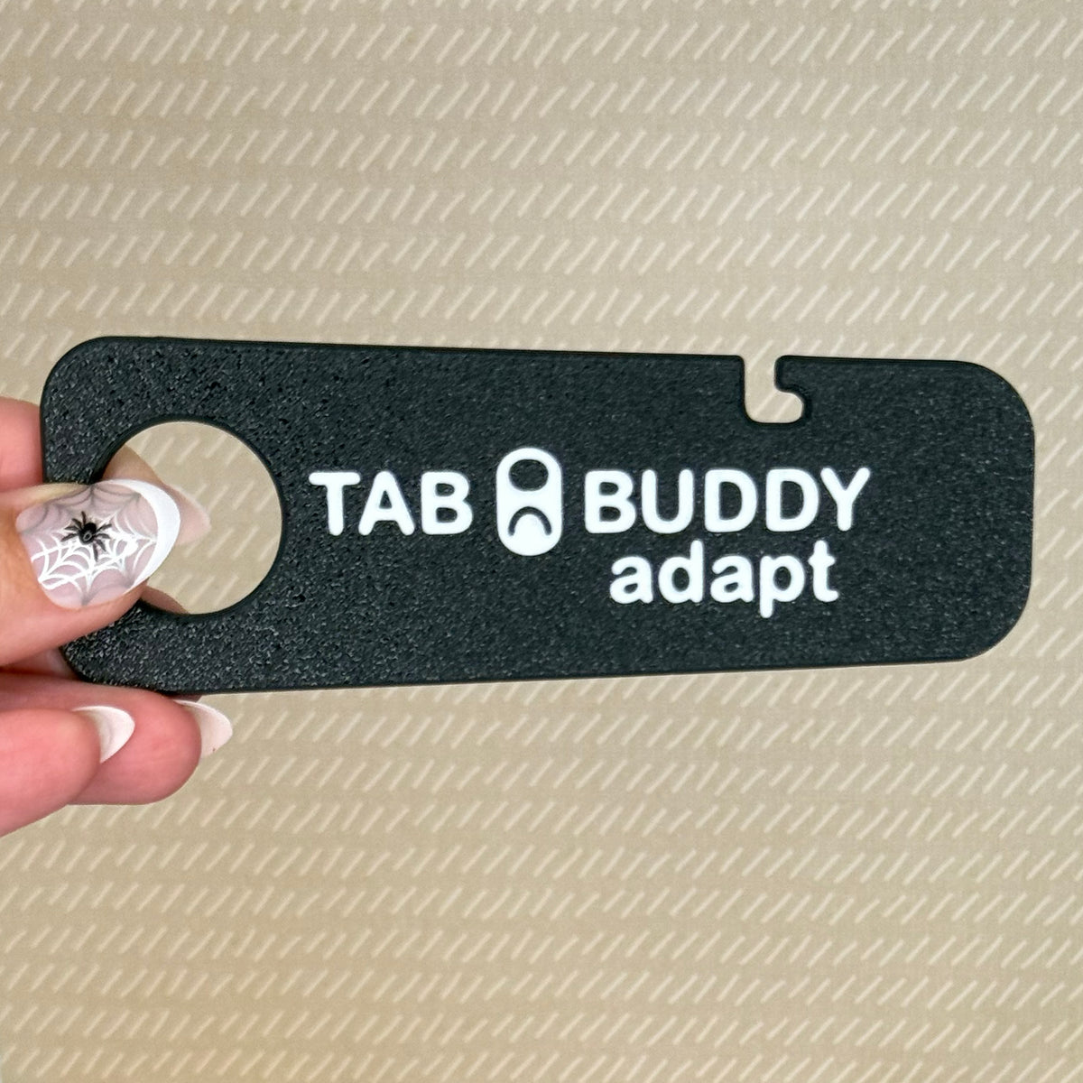 A hand with spiderweb-themed nail art holds a black Tab Buddy Adapt against a beige background.