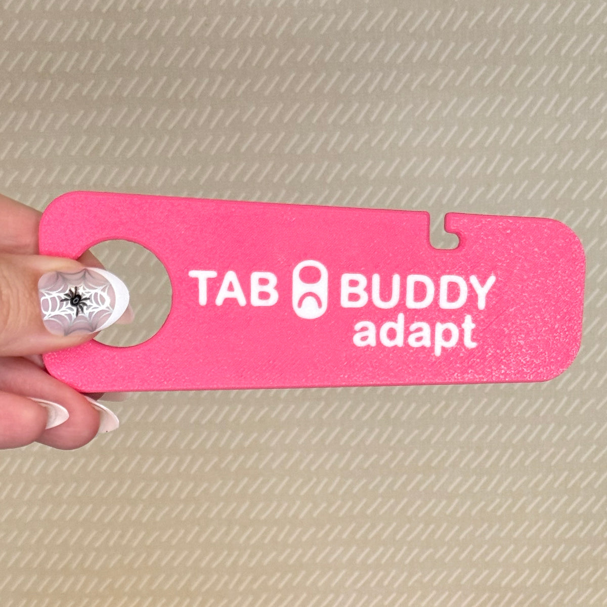 A hand with spiderweb-themed nail art holds a bright pink Tab Buddy Adapt against a beige background.