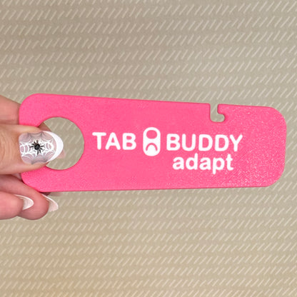 A hand with spiderweb-themed nail art holds a bright pink Tab Buddy Adapt against a beige background.