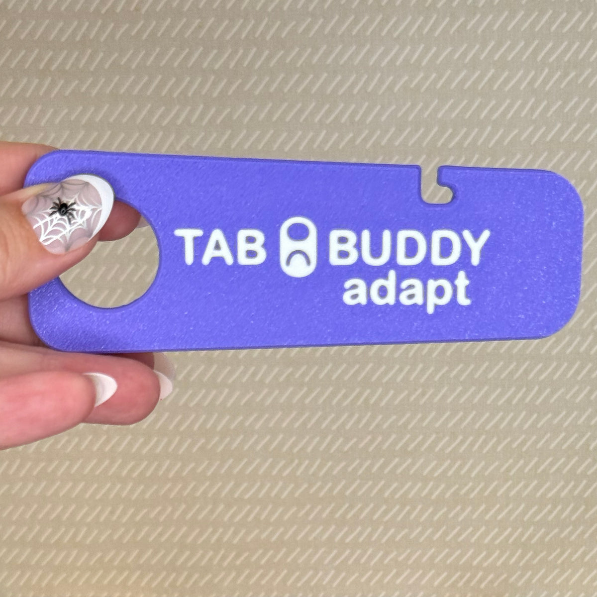 A hand with spiderweb-themed nail art holds a purple Tab Buddy Adapt against a beige background.