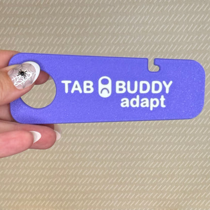 A hand with spiderweb-themed nail art holds a purple Tab Buddy Adapt against a beige background.
