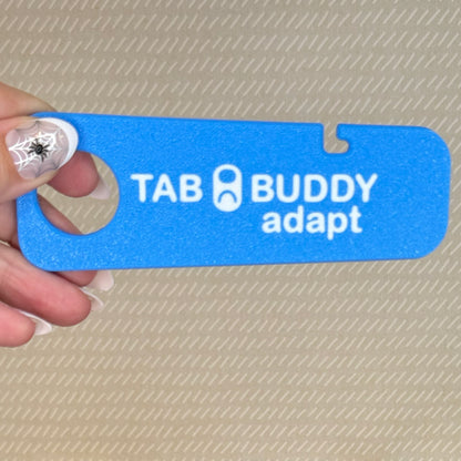 A hand with spiderweb-themed manicured nails holds a sky blue Tab Buddy Adapt against a beige background.