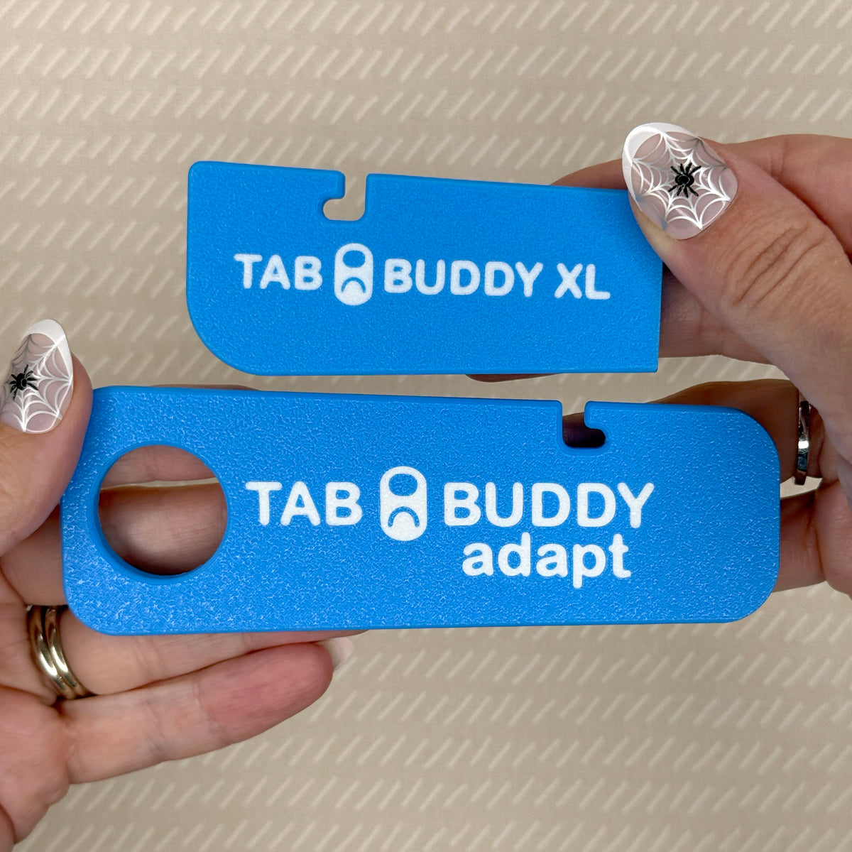 A sky blue Tab Buddy XL and Tab Buddy Adapt are shown side-by-side, emphasizing their size difference. The XL is 3 1/4 inches long, while the Adapt is 5 1/8 inches long and includes a grip assist hole.