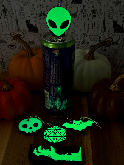 A glow-in-the-dark Soda Tab Buddy rests atop a canned drink with other glowing Tab Buddies including a skull, bat, D20 and zombine hand, resting on a wooden surface with pumpkins and a black-and-white patterned backdrop. 