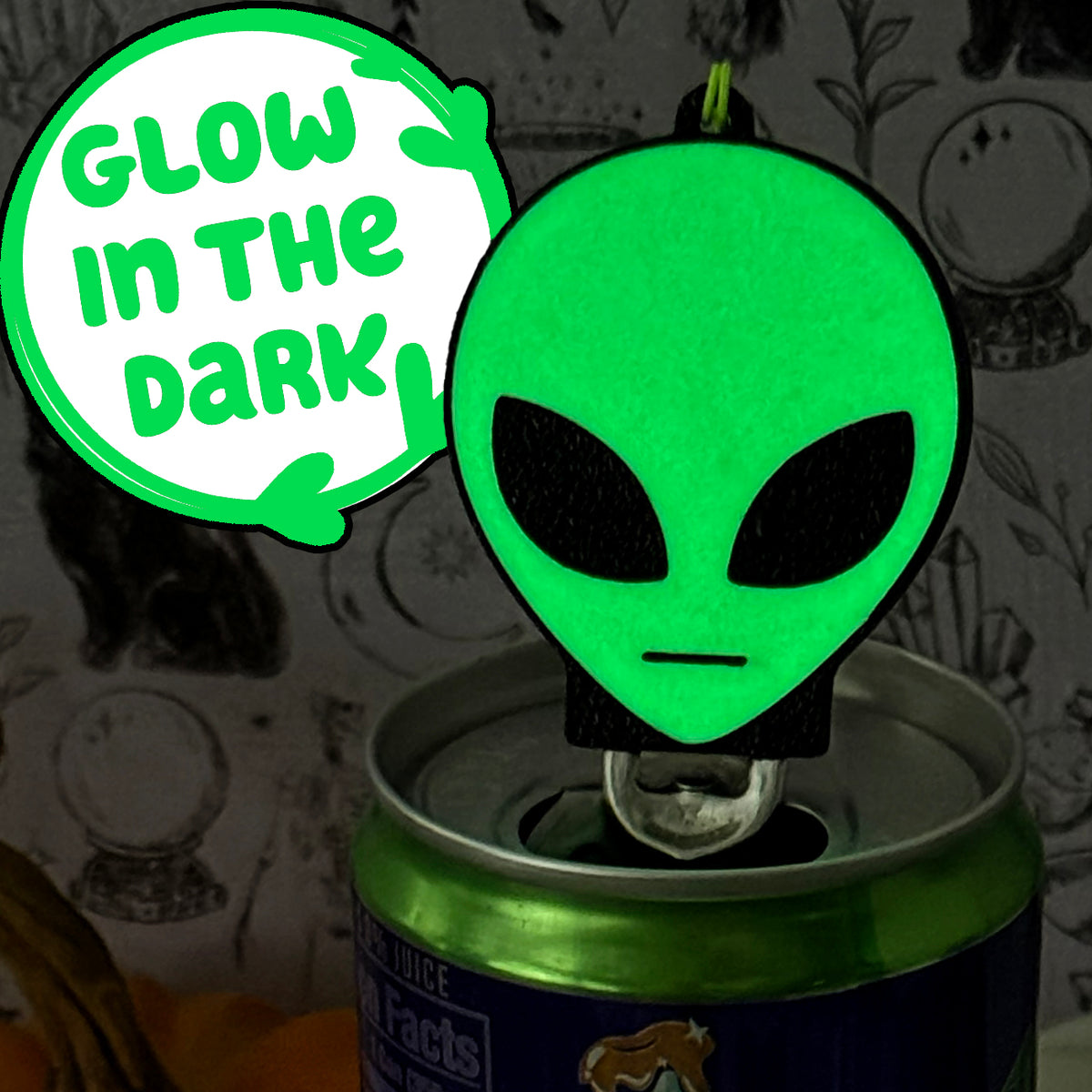 A glowing alien head Soda Tab Buddy rests atop a canned drink that has been popped open. Text bubble reads "Glow in the Dark."