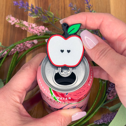 Person using a cute apple-shaped Tab Buddy on a pink sparkling water can with flowers in the background.