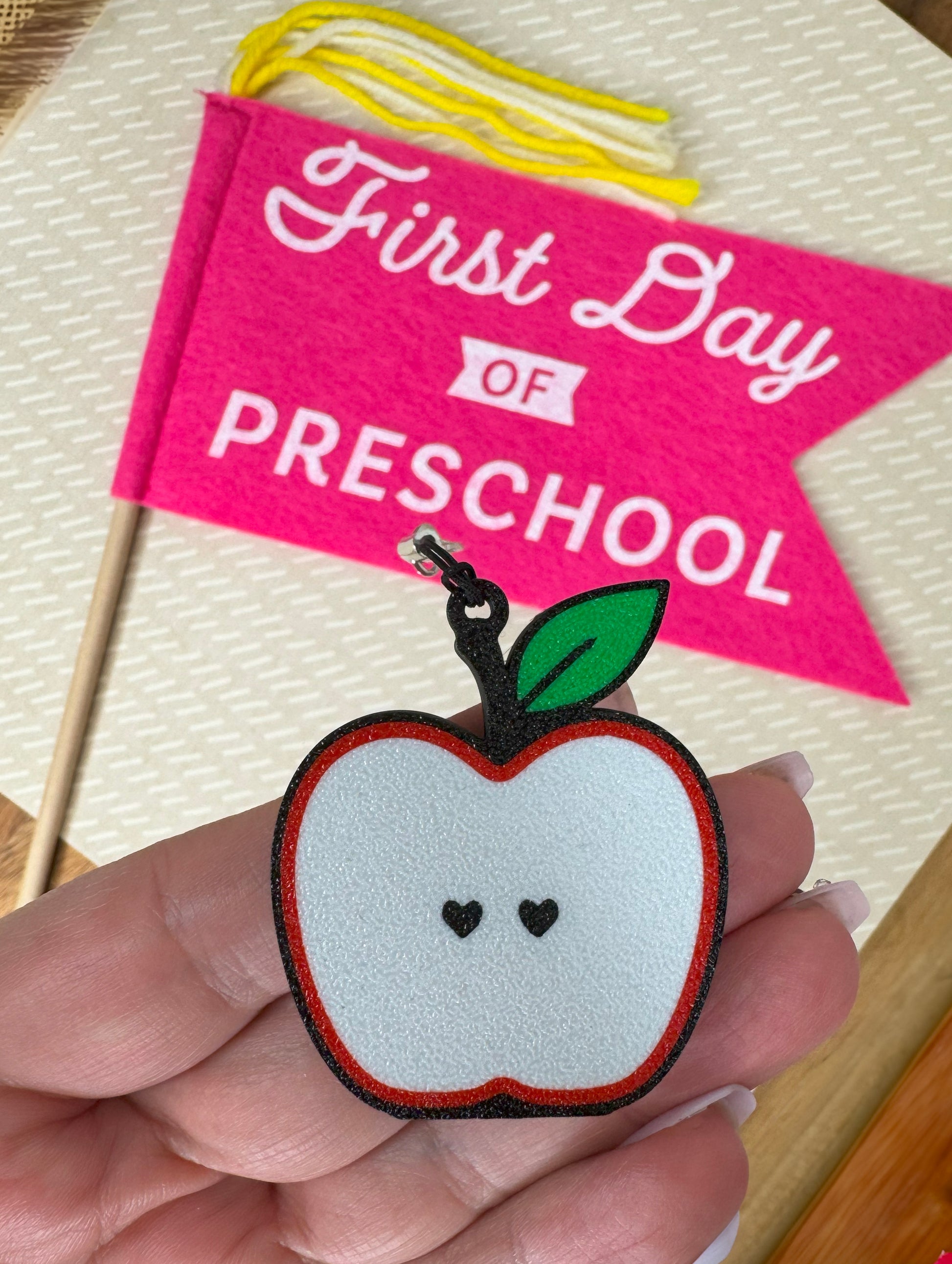Apple-shaped Tab Buddy keychain with held in front of a 'First Day of Preschool' flag.