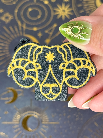 Decorative black and gold Aries Tab Buddy held by a hand with green nail polish against an astrological background.