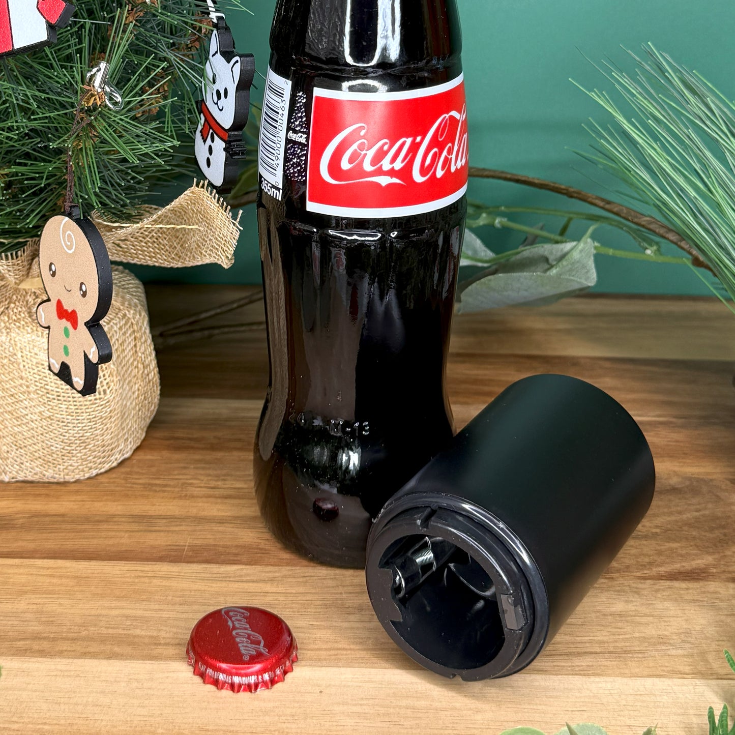 Coca-Cola bottle with cap and opener on a wooden surface with Christmas decorations in the background.