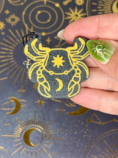 Hand holding a gold and black astrological Cancer Tab Buddy against a patterned background