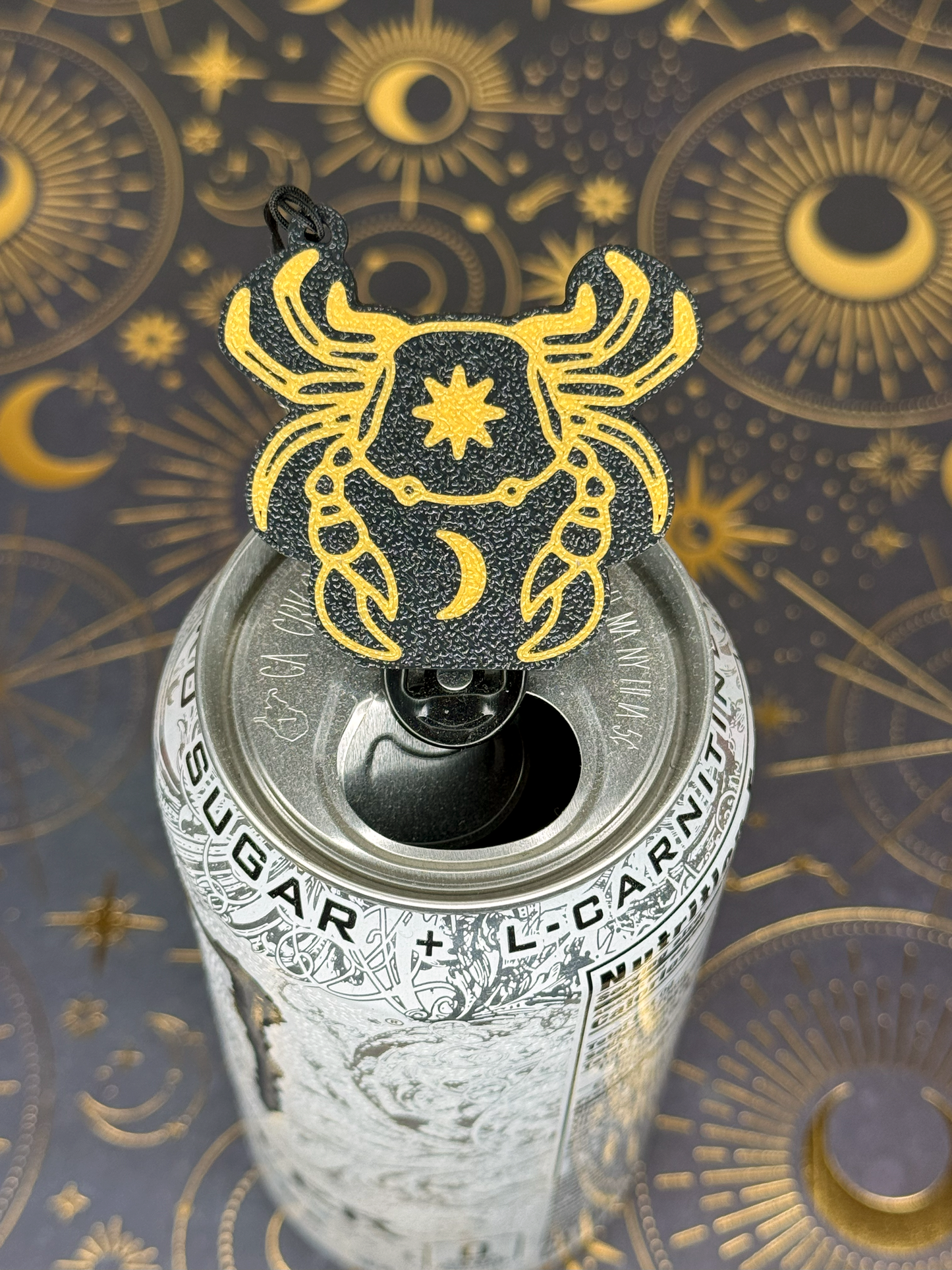 Decorative Tab Buddy keychain with a Cancer Zodiac crab star and moon design on top of a can against a celestial pattern background.