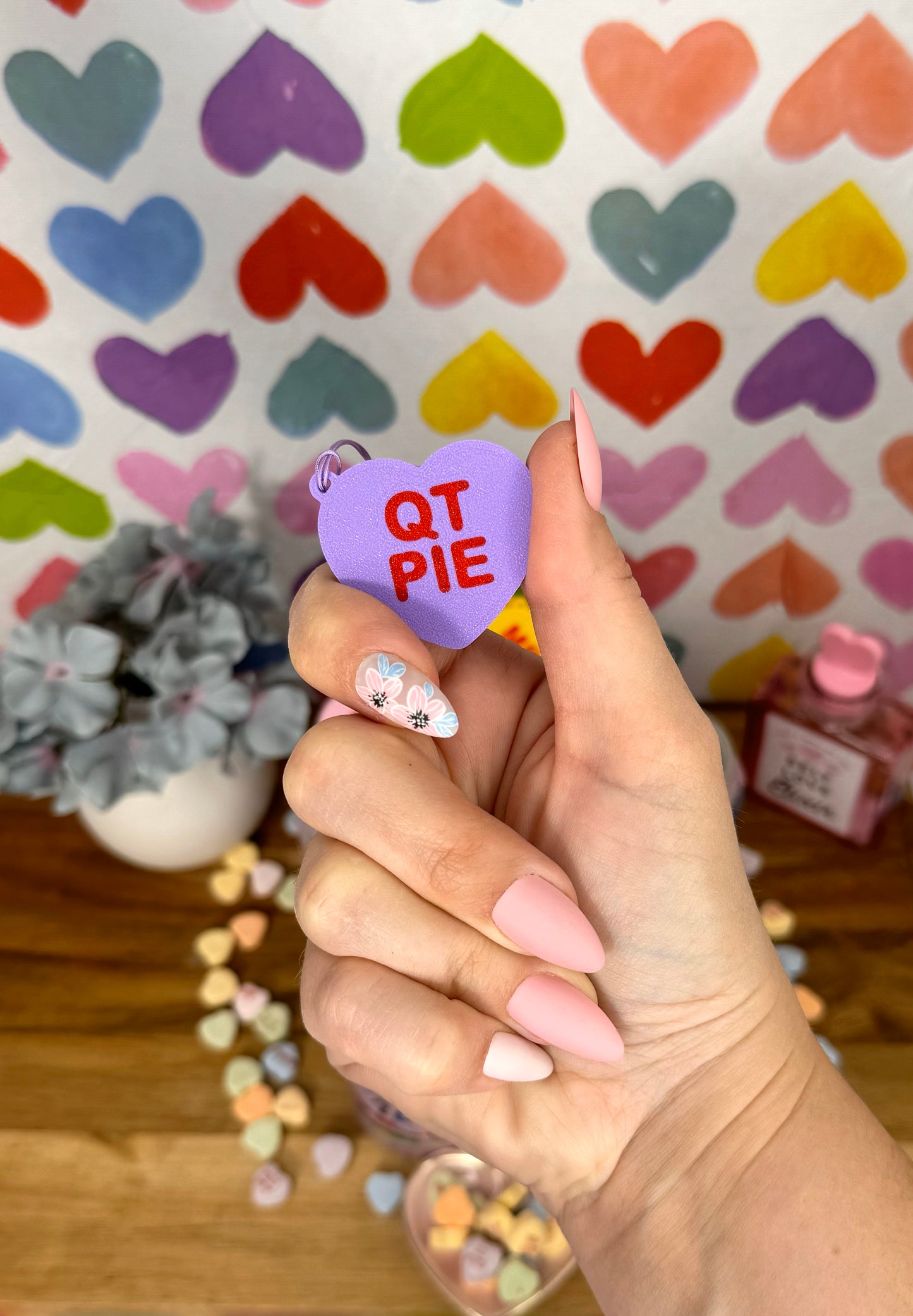 Hand holding a purple heart-shaped Tab Buddy keychain with 'QT PIE' text against a colorful heart-patterned background.