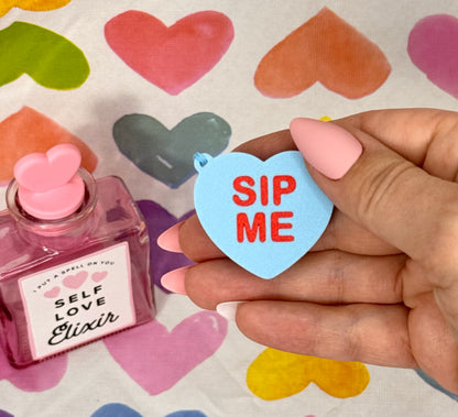 Hand holding a heart-shaped 'Sip Me' Tab Buddy next to a bottle labeled 'Self Love Elixir' on a colorful heart-patterned background.