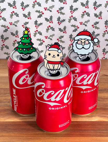 Three Coca-Cola cans with Christmas-themed Tab Buddies on a wooden surface with a holly leaf pattern.