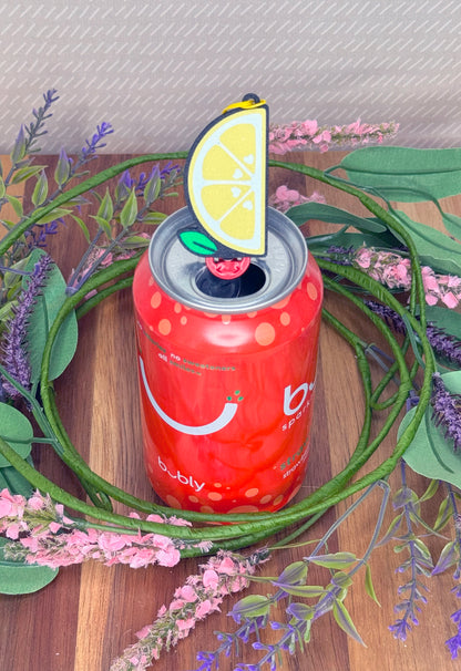 Red can of Bubly with a lemon slice Tab Buddy on top, surrounded by green leaves and pink flowers.