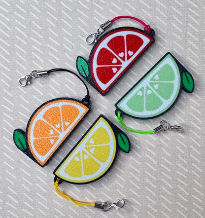 Four colorful citrus-shaped Tab Buddy keychains on a textured beige background