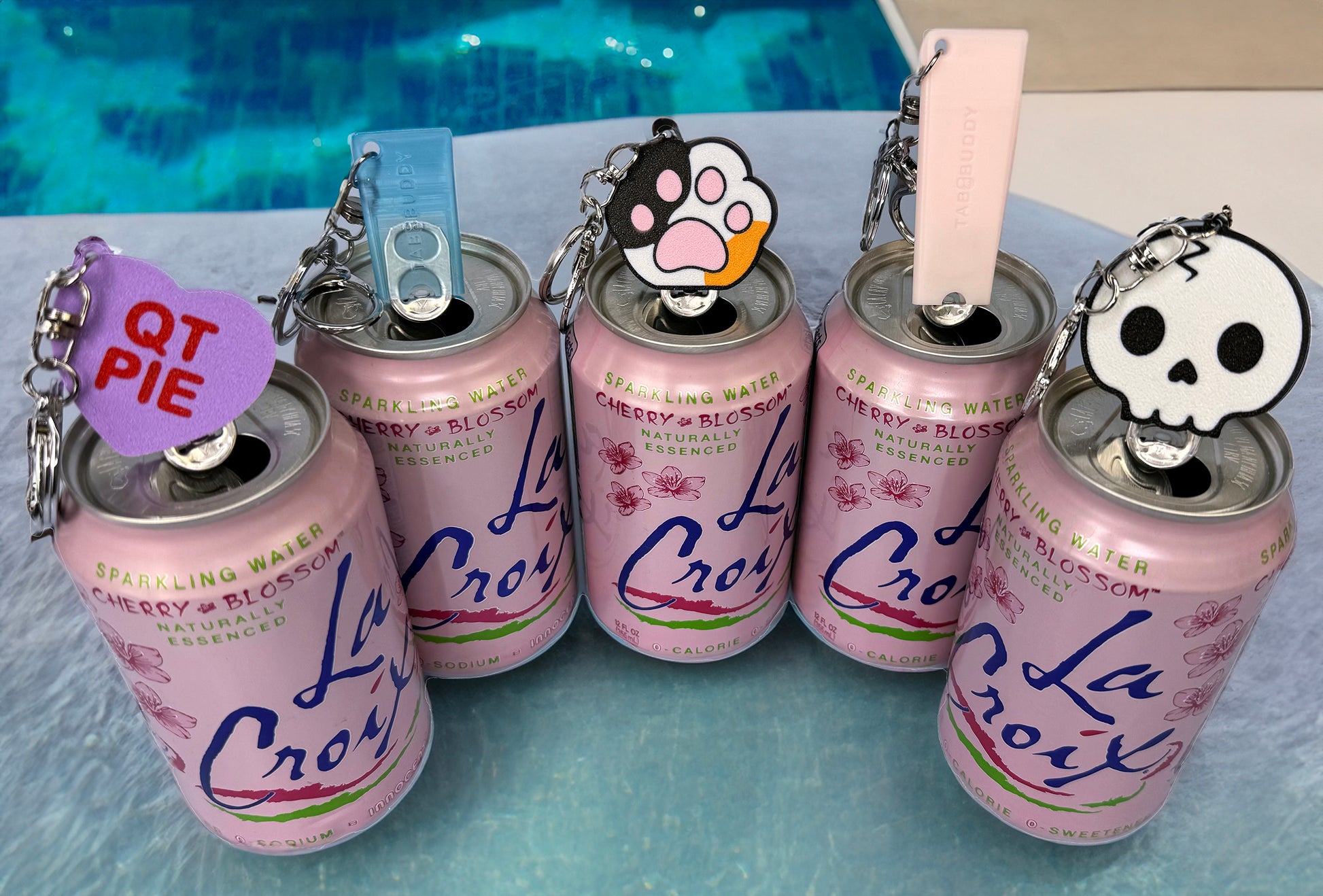 Five pink La Croix cans with various Tab Buddies shown with optional add-on keychains on a blue surface.
