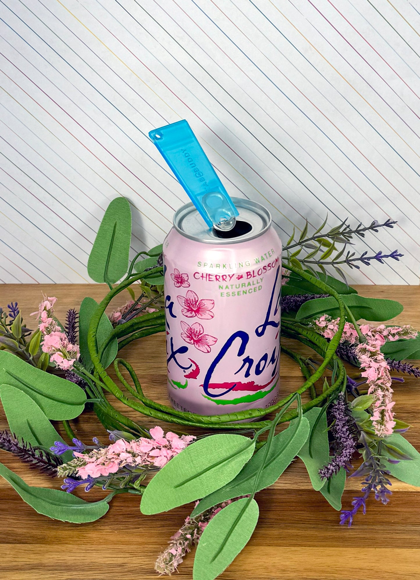 Pink can of La Croix with a Sapphire blue Tab Buddy Classic on a wooden surface with flowers and leaves.