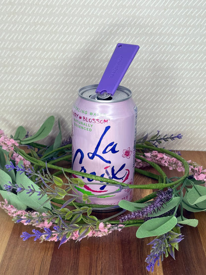A purple Tab Buddy Classic soda can opener rests atop the tab of an open can of sparkling water with a beige background and floral accents.