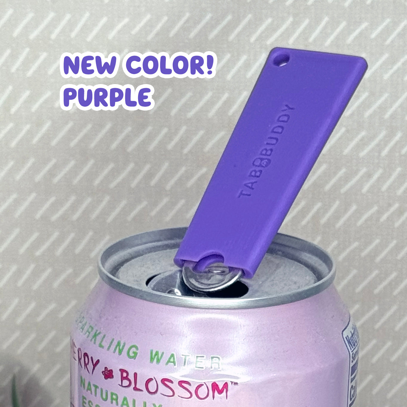 A purple Tab Buddy Classic soda can opener rests atop the tab of an open can of sparkling water with a beige background. Text reads "New Color! Purple"