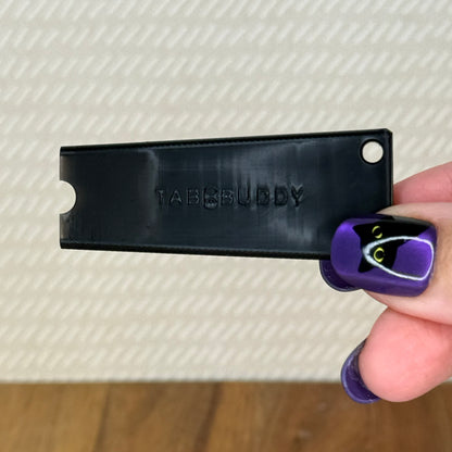 A black Tab Buddy Classic is held by a hand with purple and black nail art against a textured beige background.