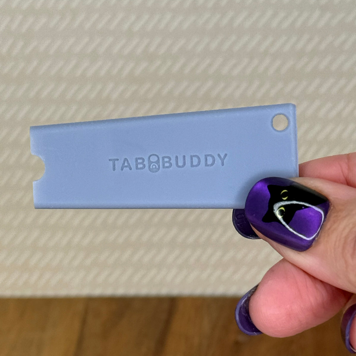 A grey Tab Buddy Classic is held by a hand with purple and black nail art against a textured beige background.