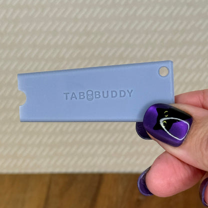 A grey Tab Buddy Classic is held by a hand with purple and black nail art against a textured beige background.