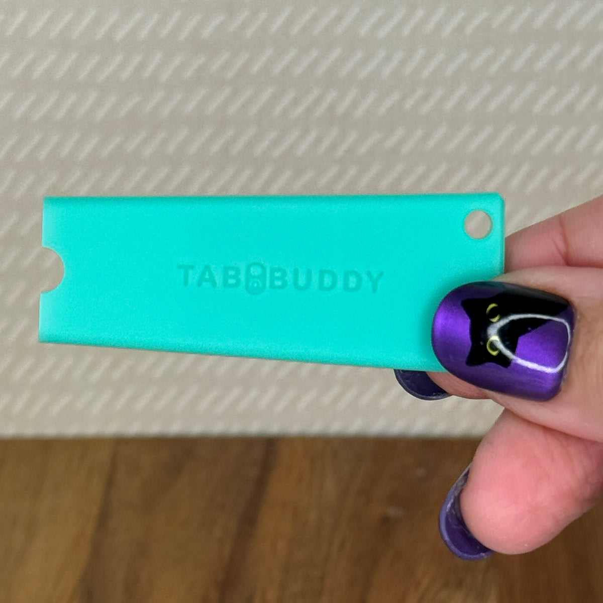 A jade Tab Buddy Classic is held by a hand with purple and black nail art against a textured beige background.