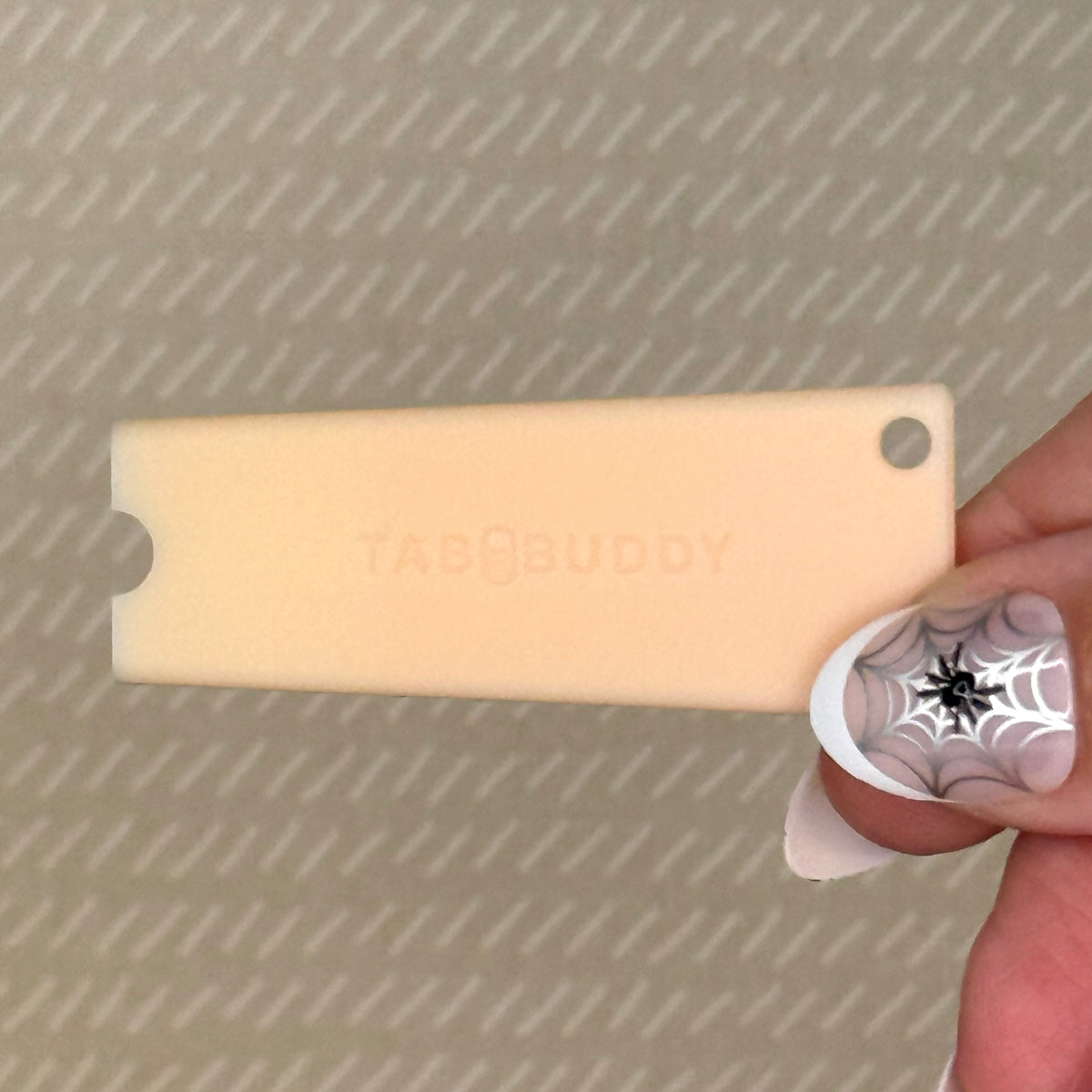 A peach Tab Buddy Classic is held by a hand with purple and black nail art against a textured beige background.
