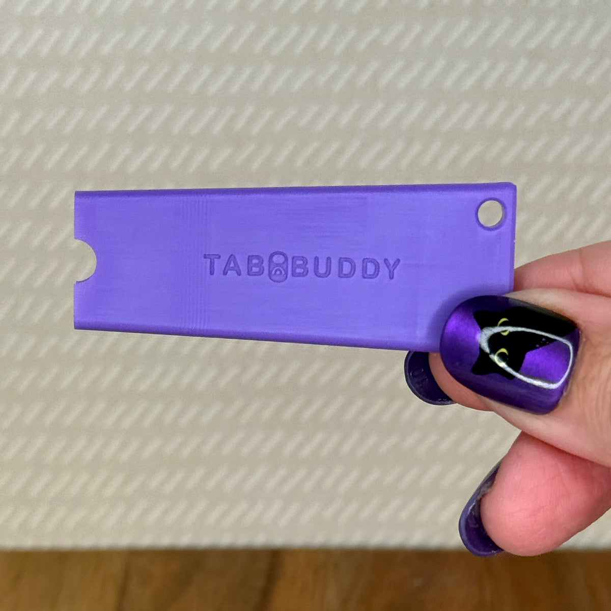 A purple Tab Buddy Classic is held by a hand with purple and black nail art against a textured beige background.
