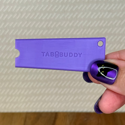 A purple Tab Buddy Classic is held by a hand with purple and black nail art against a textured beige background.