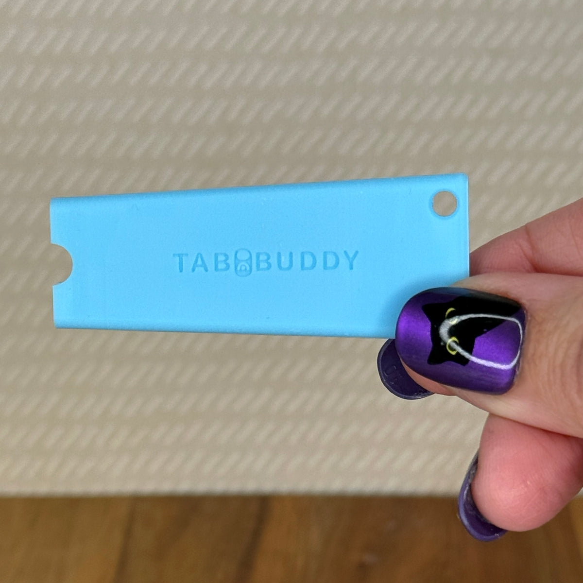 A sky blue Tab Buddy Classic is held by a hand with purple and black nail art against a textured beige background.
