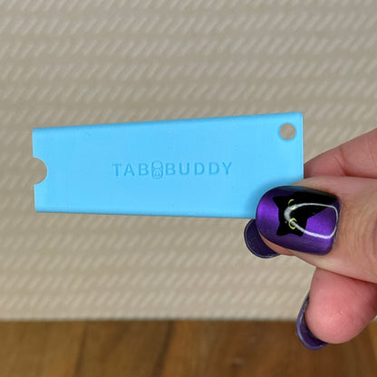 A sky blue Tab Buddy Classic is held by a hand with purple and black nail art against a textured beige background.