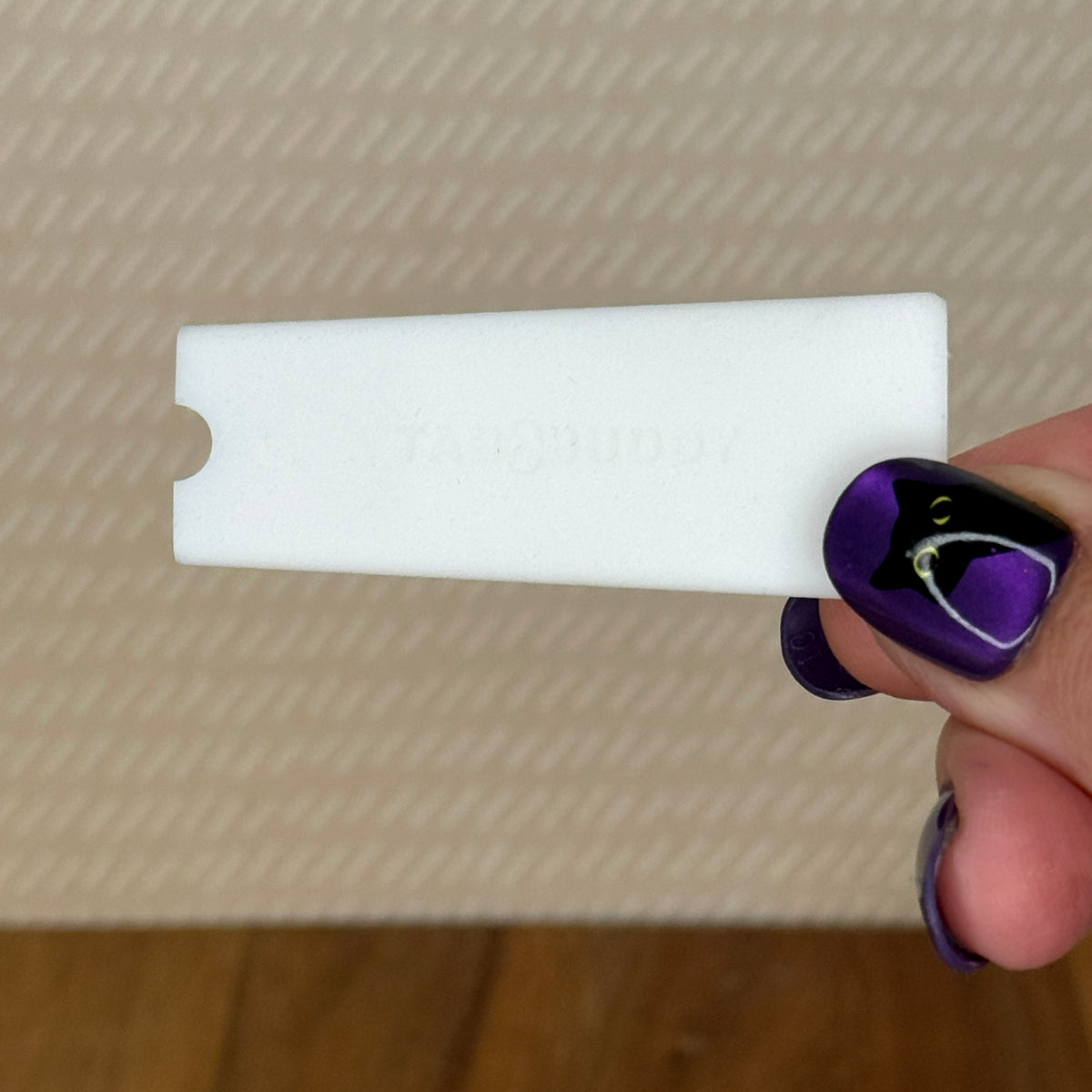 A white Tab Buddy Classic is held by a hand with purple and black nail art against a textured beige background.