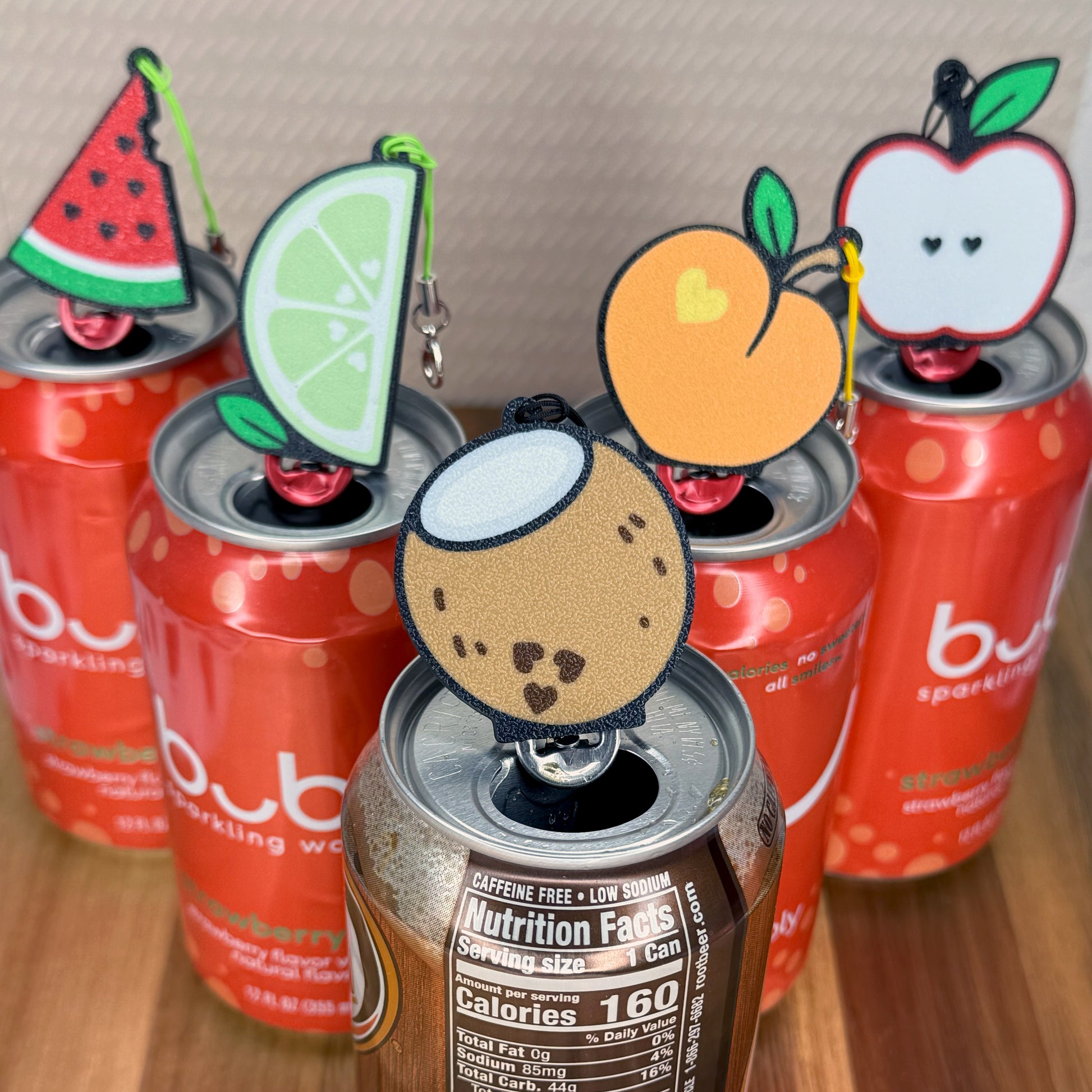 Red cans with fruit-themed Tab Buddies on a wooden surface