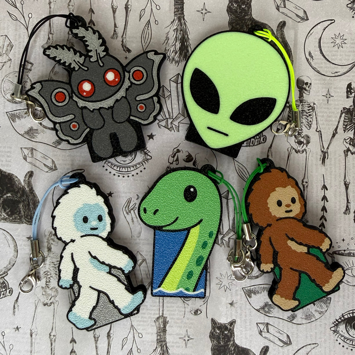 A group of 5 cryptid-themed Soda Tab Buddies rest on a patterned grey background as part of the 2026 Halloween Collection. They include Mothman, an Alien head, a Yeti, a Loch Ness Monster, and a Bigfoot. Each one has a keychain strap attached to it.