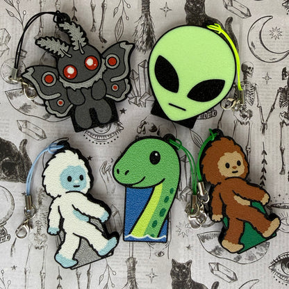 A group of 5 cryptid-themed Soda Tab Buddies rest on a patterned grey background as part of the 2026 Halloween Collection. They include Mothman, an Alien head, a Yeti, a Loch Ness Monster, and a Bigfoot. Each one has a keychain strap attached to it.