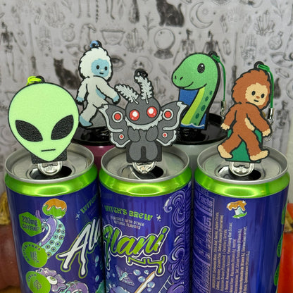 A group of 5 cryptid-themed Soda Tab Buddies rest on top of the tabs of open cans as part of the 2026 Halloween Collection. They include Mothman, an Alien head, a Yeti, a Loch Ness Monster, and a Bigfoot. Each one has a keychain strap attached to it. A patterned grey wallpaper is in the  background.