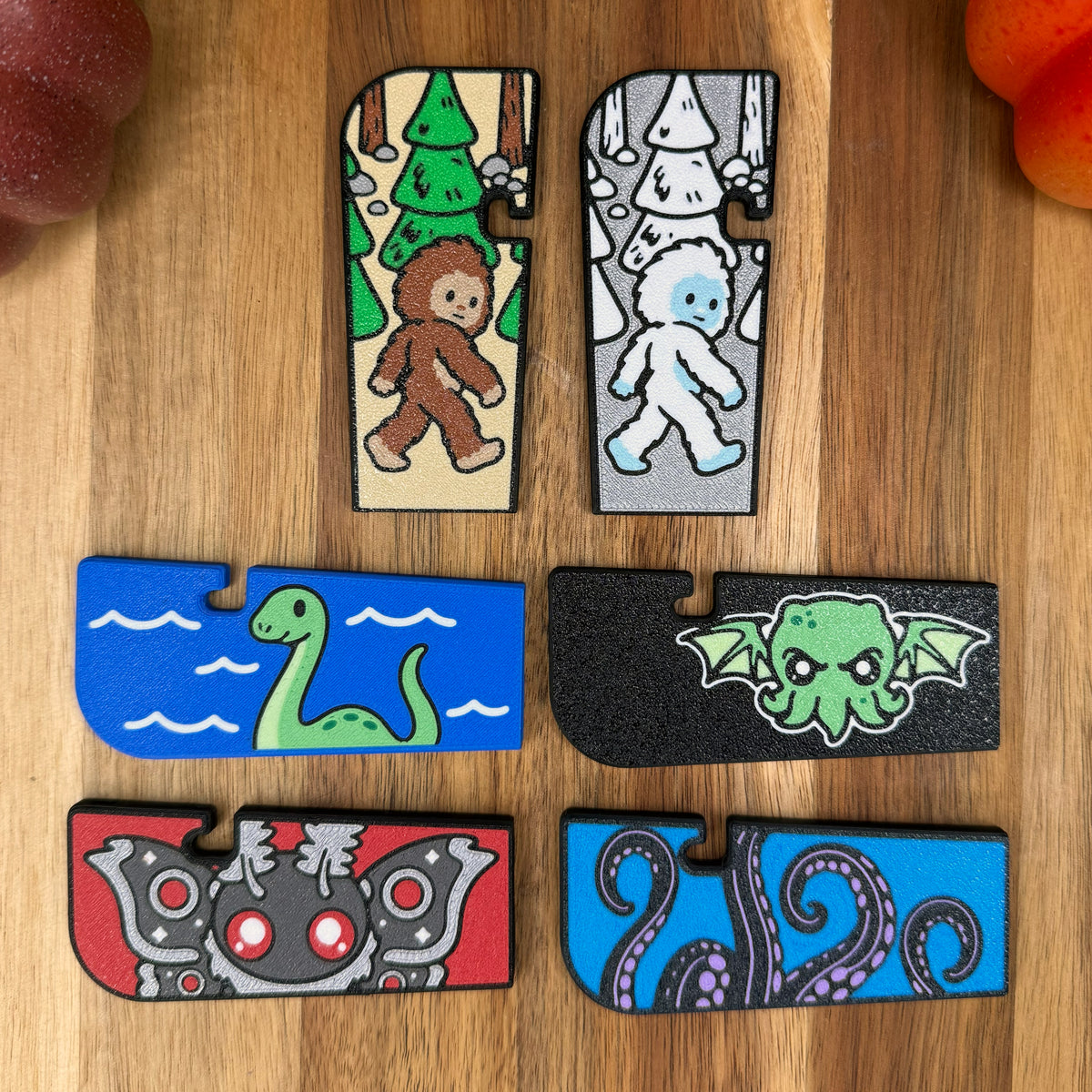 A set of 6 crytid-themed Tab Buddy XLs rest atop a wooden surface. The designs include a brown and tan Bigfoot, silver and white snow yeti, blue and green Loch Ness Monster, black and green Cthulhu. red and grey Mothman, and blue and purple Kraken.