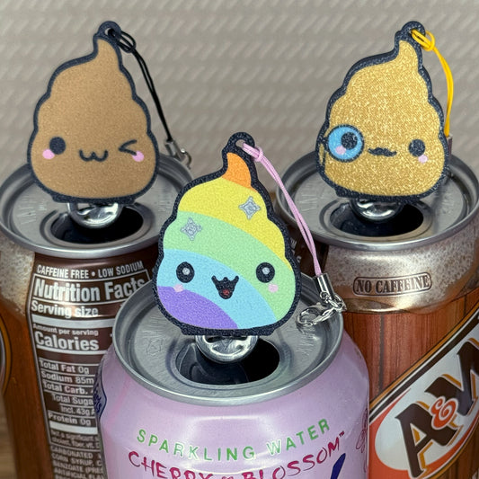 Three cute poop emoji-shaped Tab Buddy keychains on top of soda cans. ne brown, one gold, one rainbow.