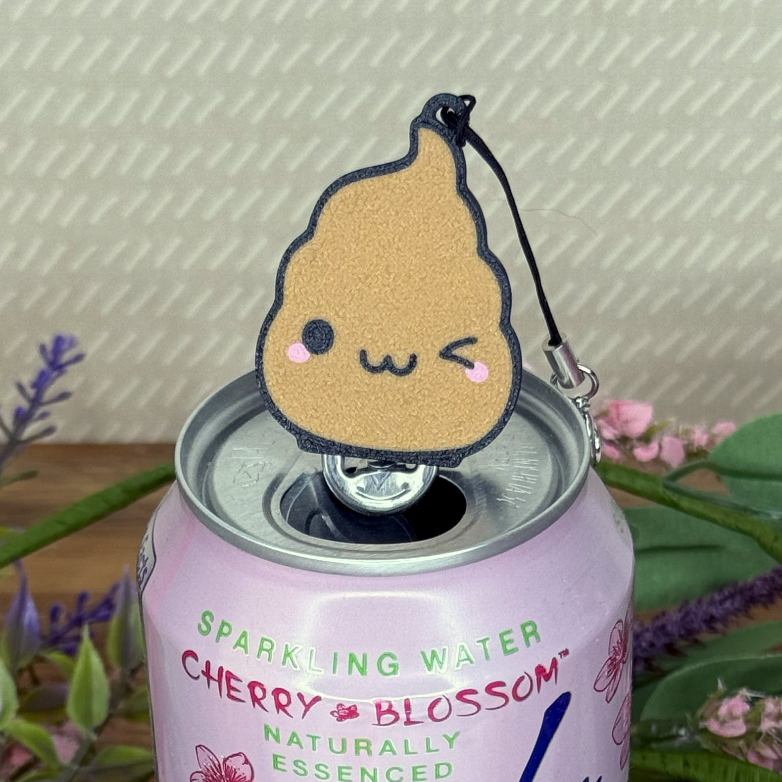 Brown poop-shaped Tab Buddy keychain with a winking smiley face sits atop a cherry blossom sparkling water can with a floral background