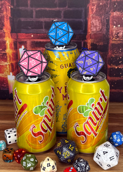 Three squirt soda cans with D20 dice Tab Buddies on top against a brick wall background