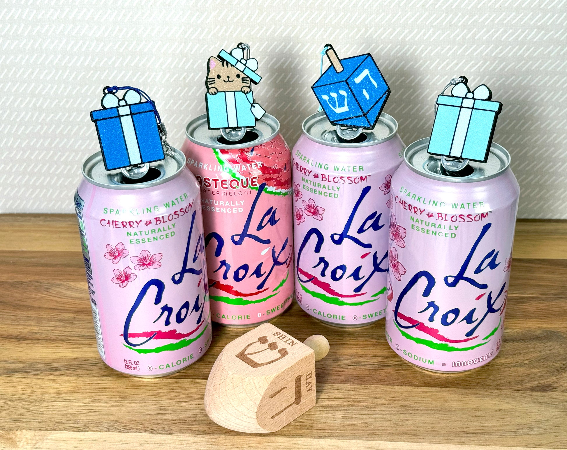 Four La Croix cans with Tab Buddies in different shades of blue resting atop the tabs on a wooden surface with a wooden dreidel displaying "shin" and "hay" on it.